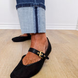 Black flat shoes with a strap worn with rolled-up jeans on a wooden floor.
