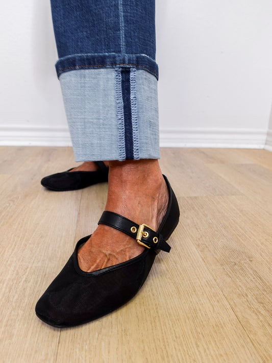 Black flat shoes with a strap worn with rolled-up jeans on a wooden floor.
