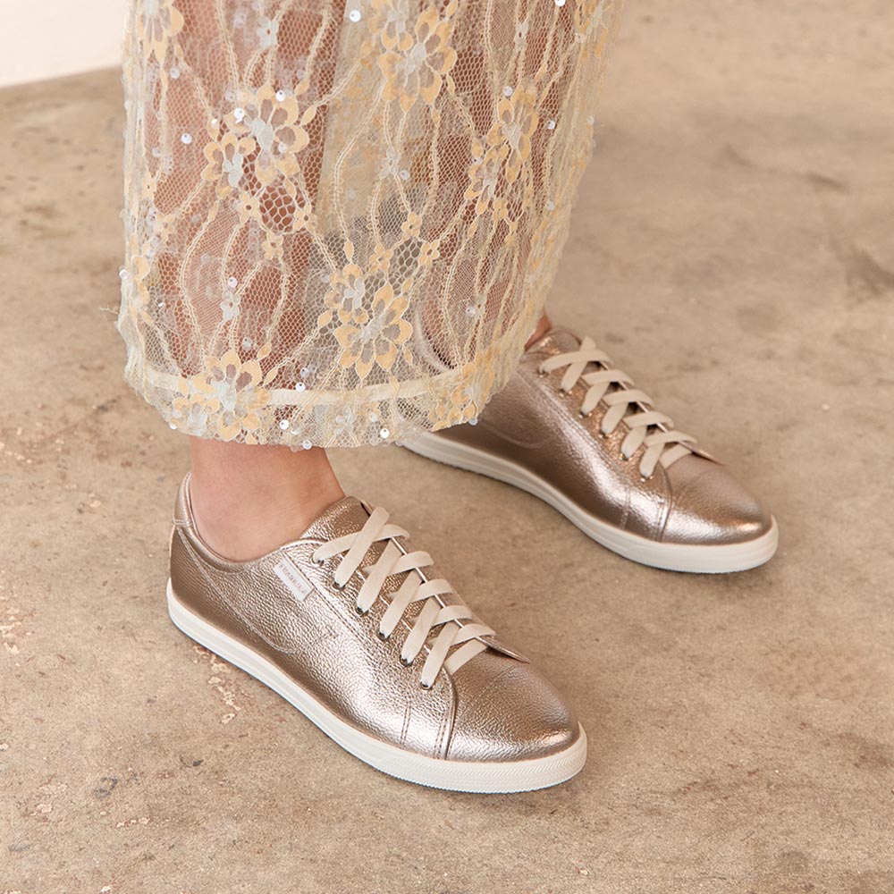 Bronze sneakers worn with sheer lace pants on a neutral background