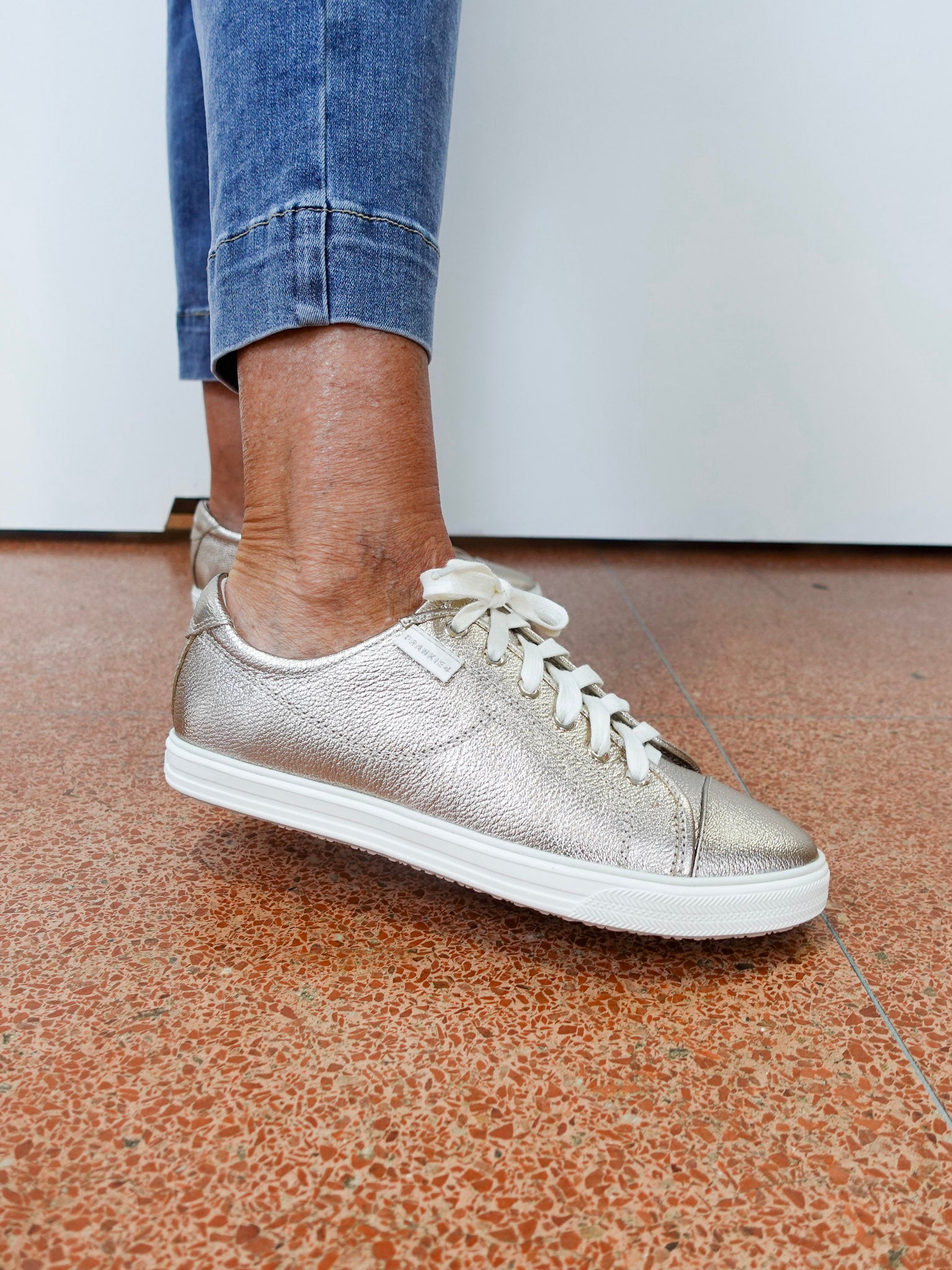 Silver sneaker worn with blue jeans on a brown floor.