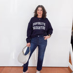 Woman wearing a navy sweatshirt with 'FAVORITE DAUGHTER' text, holding a white handbag against a white wall.
