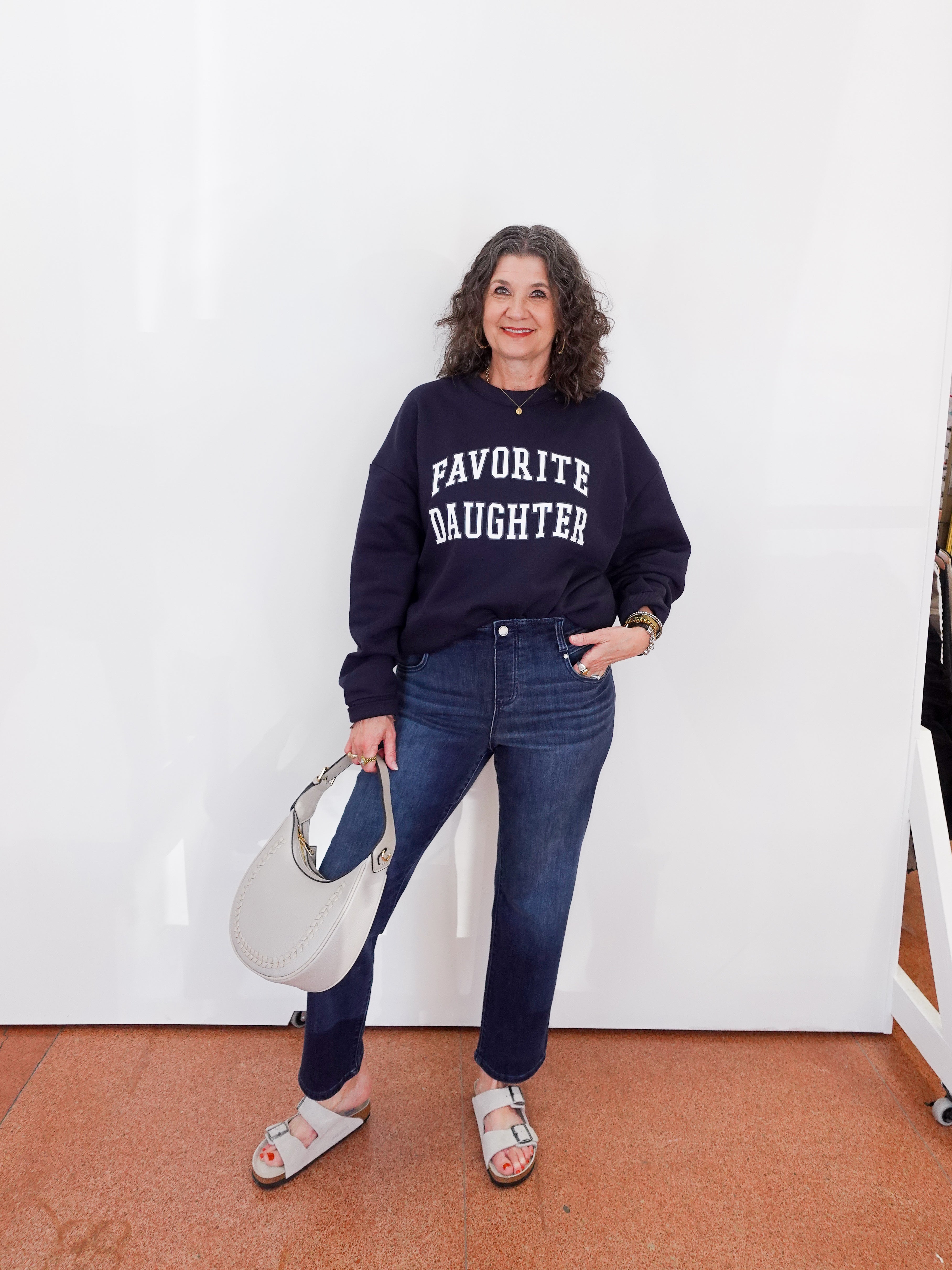 Woman wearing a navy sweatshirt with 'FAVORITE DAUGHTER' text, holding a white handbag against a white wall.