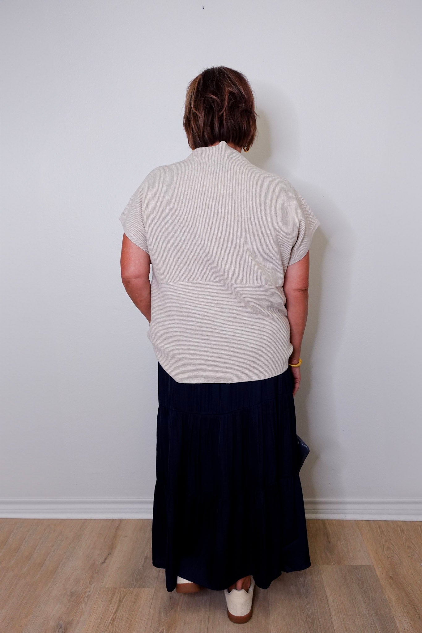 Person wearing a light gray sleeveless top and dark blue skirt against a white wall.
