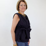 Woman wearing a black sleeveless top with a knot detail and blue jeans against a white wall.