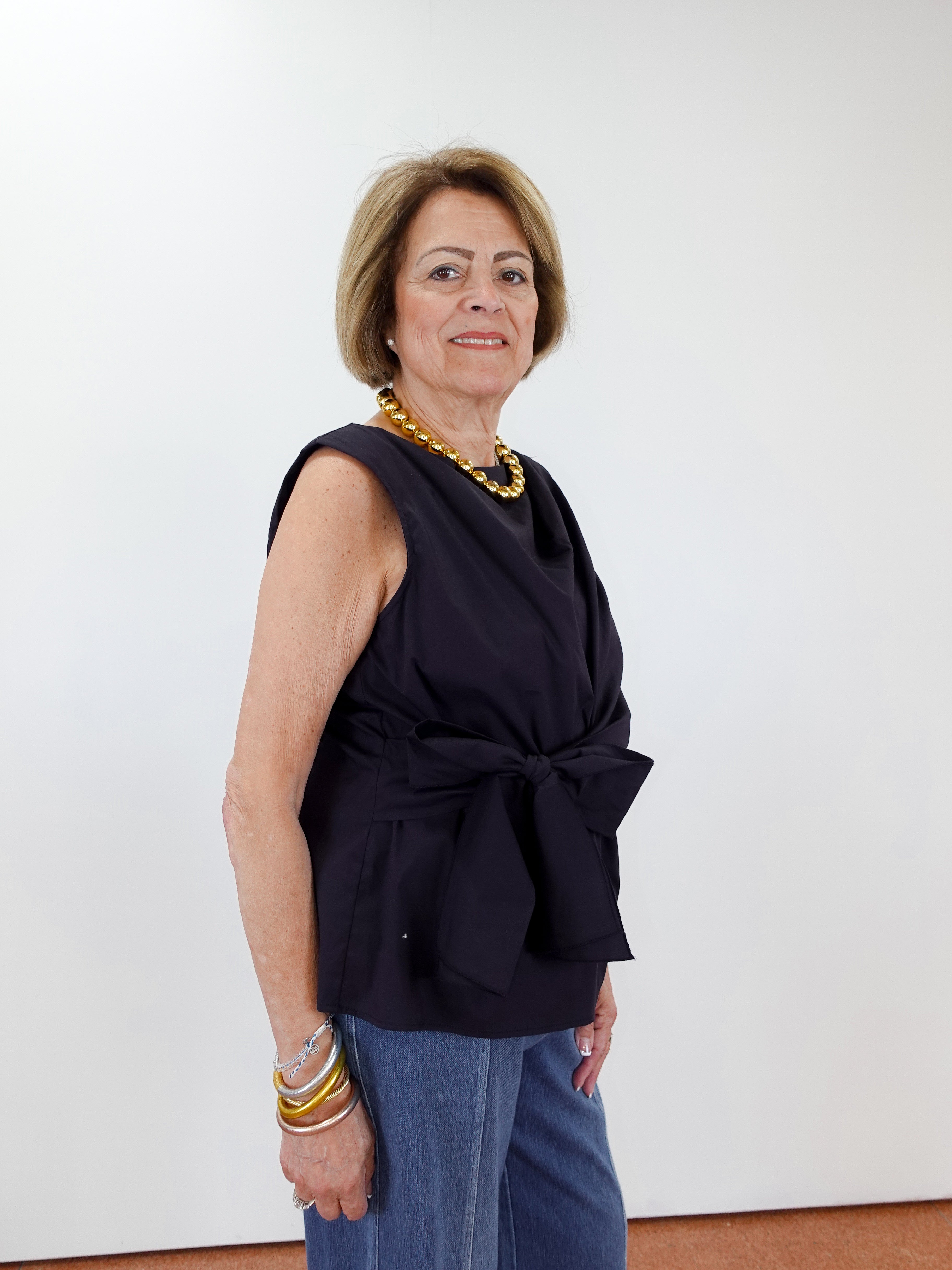 Woman wearing a black sleeveless top with a knot detail and blue jeans against a white wall.