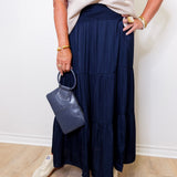 Person wearing a navy blue skirt and white top, holding a gray handbag against a white wall.