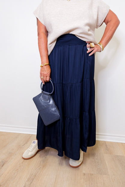 Person wearing a navy blue skirt and white top, holding a gray handbag against a white wall.