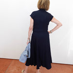 Person wearing a navy dress holding a light blue jacket against a white wall.