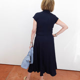 Person wearing a navy dress holding a light blue jacket against a white wall.