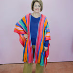 Woman wearing a colorful striped poncho over a blue top and brown pants against a pink wall.
