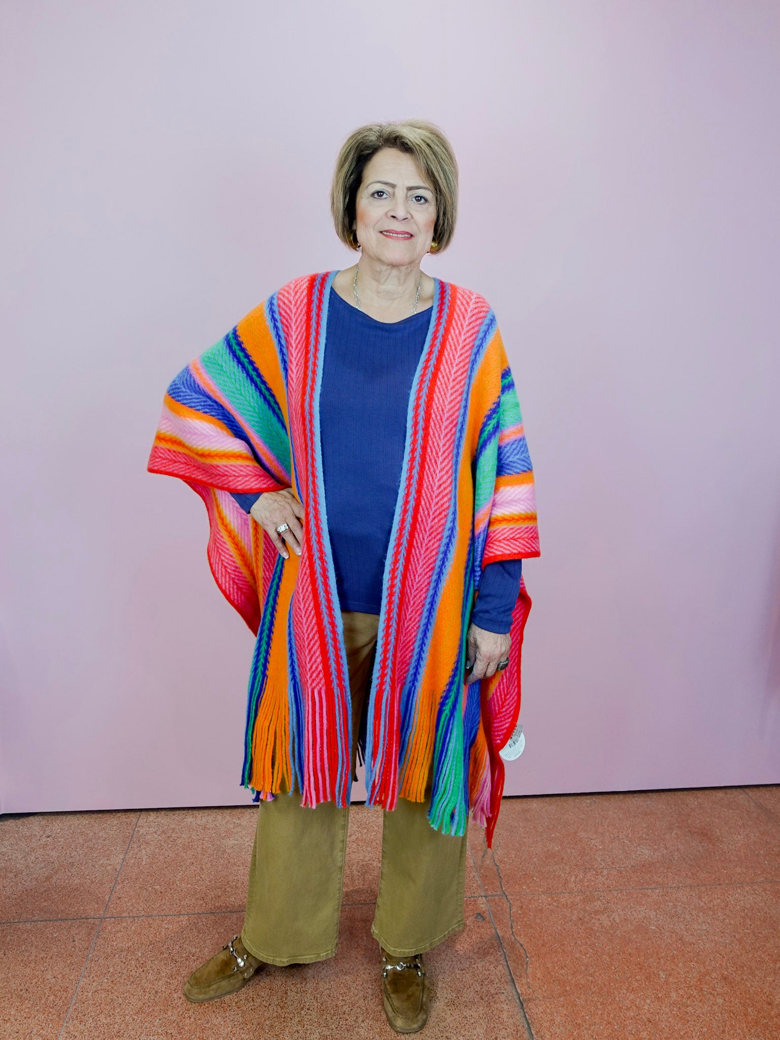 Woman wearing a colorful striped poncho over a blue top and brown pants against a pink wall.