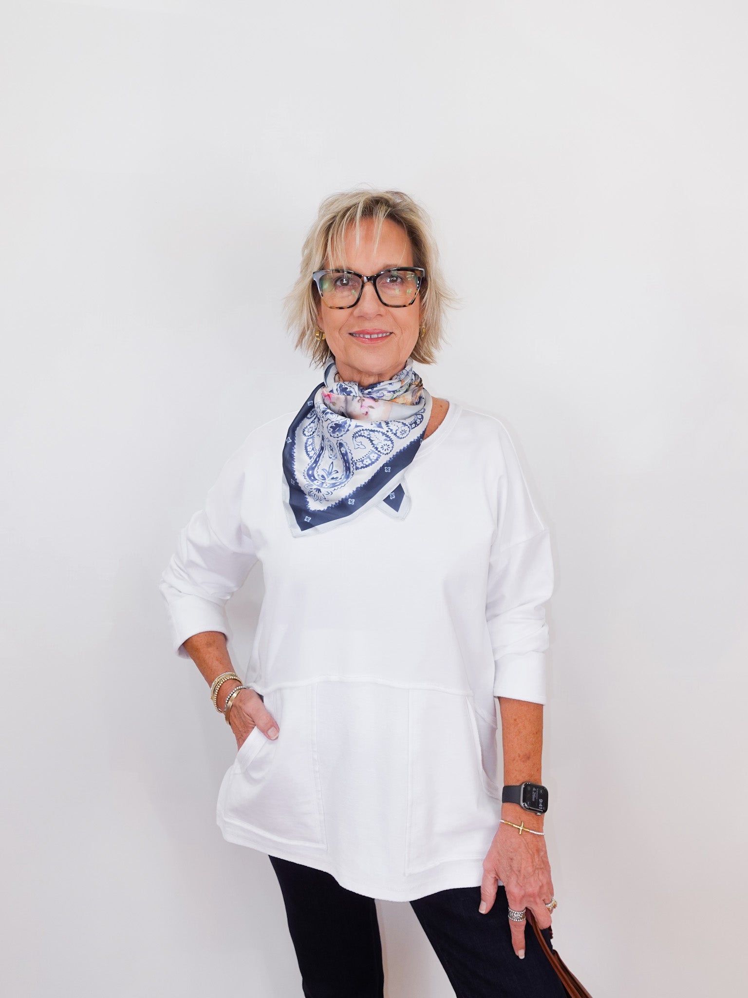 Person wearing a white shirt and black pants with a patterned scarf, standing against a plain background
