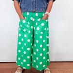 A person wearing bright green wide-leg pants with large white polka dots, paired with a blue denim shirt and beige shoes, standing against a plain white wall.