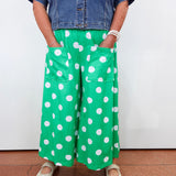 A person wearing bright green wide-leg pants with large white polka dots, paired with a blue denim shirt and beige shoes, standing against a plain white wall.