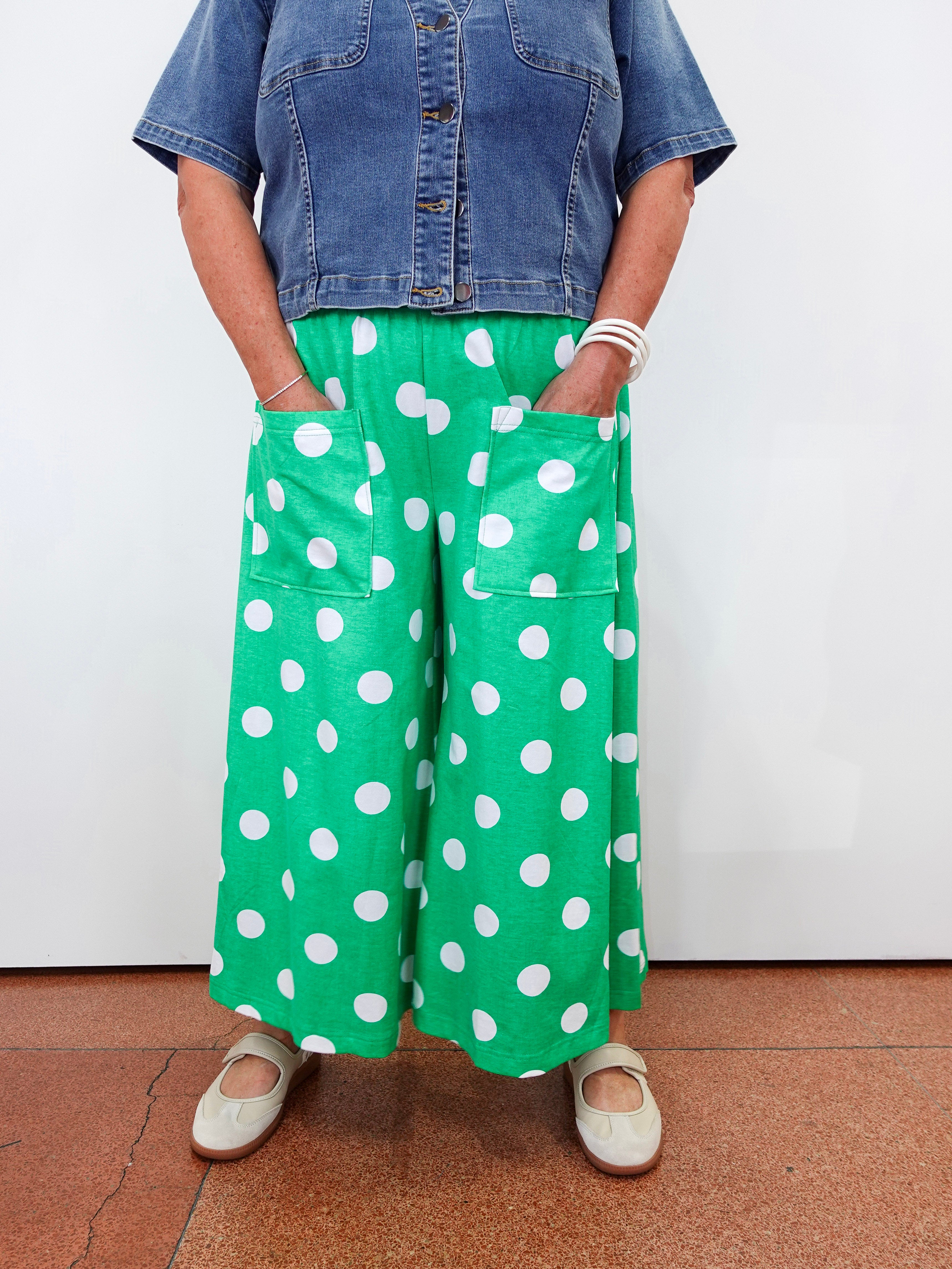 A person wearing bright green wide-leg pants with large white polka dots, paired with a blue denim shirt and beige shoes, standing against a plain white wall.