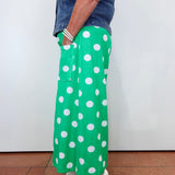 A person wearing bright green wide-leg pants with large white polka dots, paired with a denim shirt and white slide sandals, standing against a plain white wall.