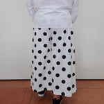 A person shown from the back wearing a white long-sleeve top and wide-leg white pants with large black polka dots, standing on a terracotta-colored floor against a plain off-white wall.