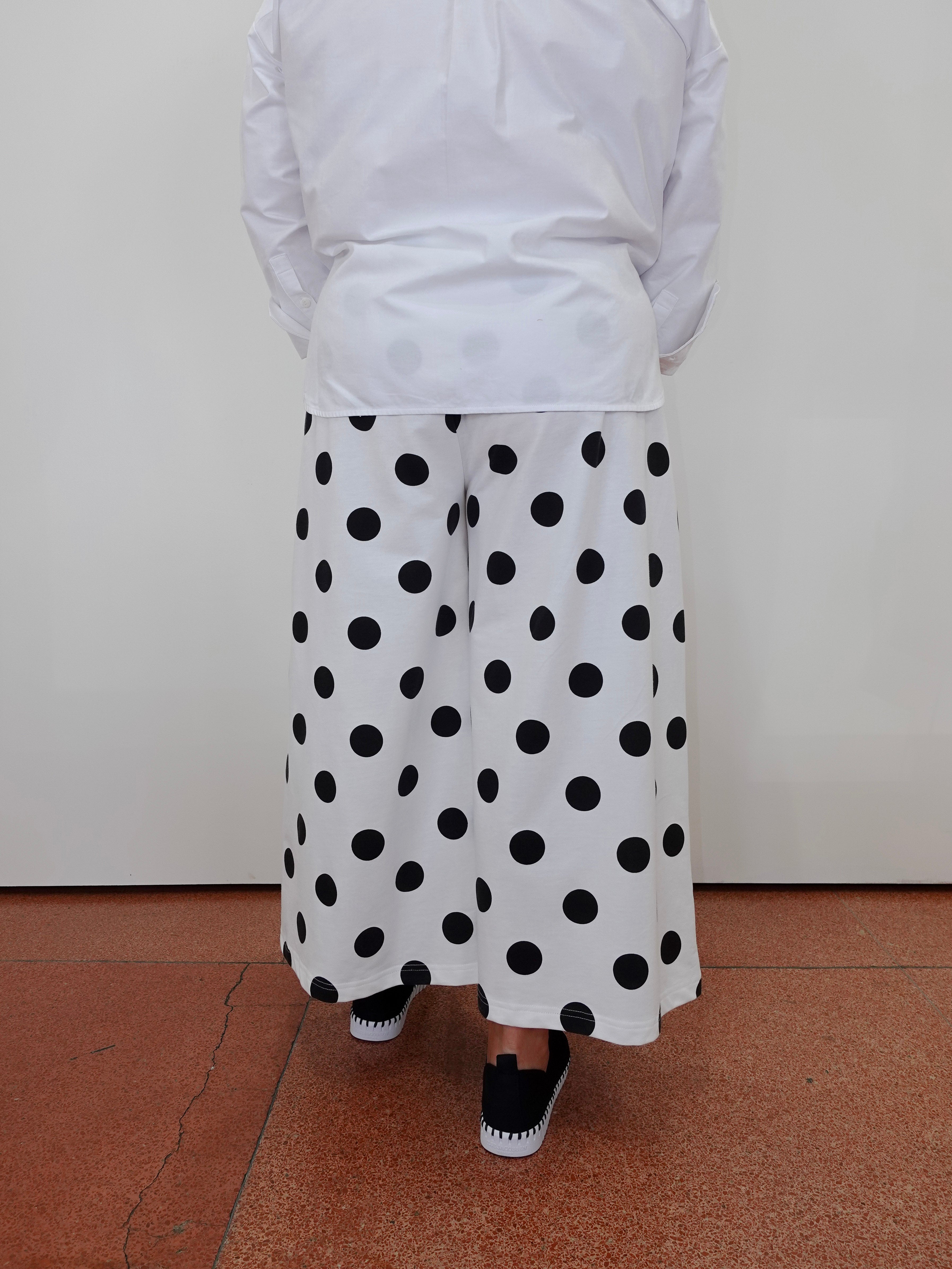 A person shown from the back wearing a white long-sleeve top and wide-leg white pants with large black polka dots, standing on a terracotta-colored floor against a plain off-white wall.