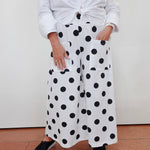 A person wearing white wide-leg pants with large black polka dots, paired with a white tied blouse and black espadrille sandals, standing against a plain off-white wall.