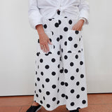 A person wearing white wide-leg pants with large black polka dots, paired with a white tied blouse and black espadrille sandals, standing against a plain off-white wall.