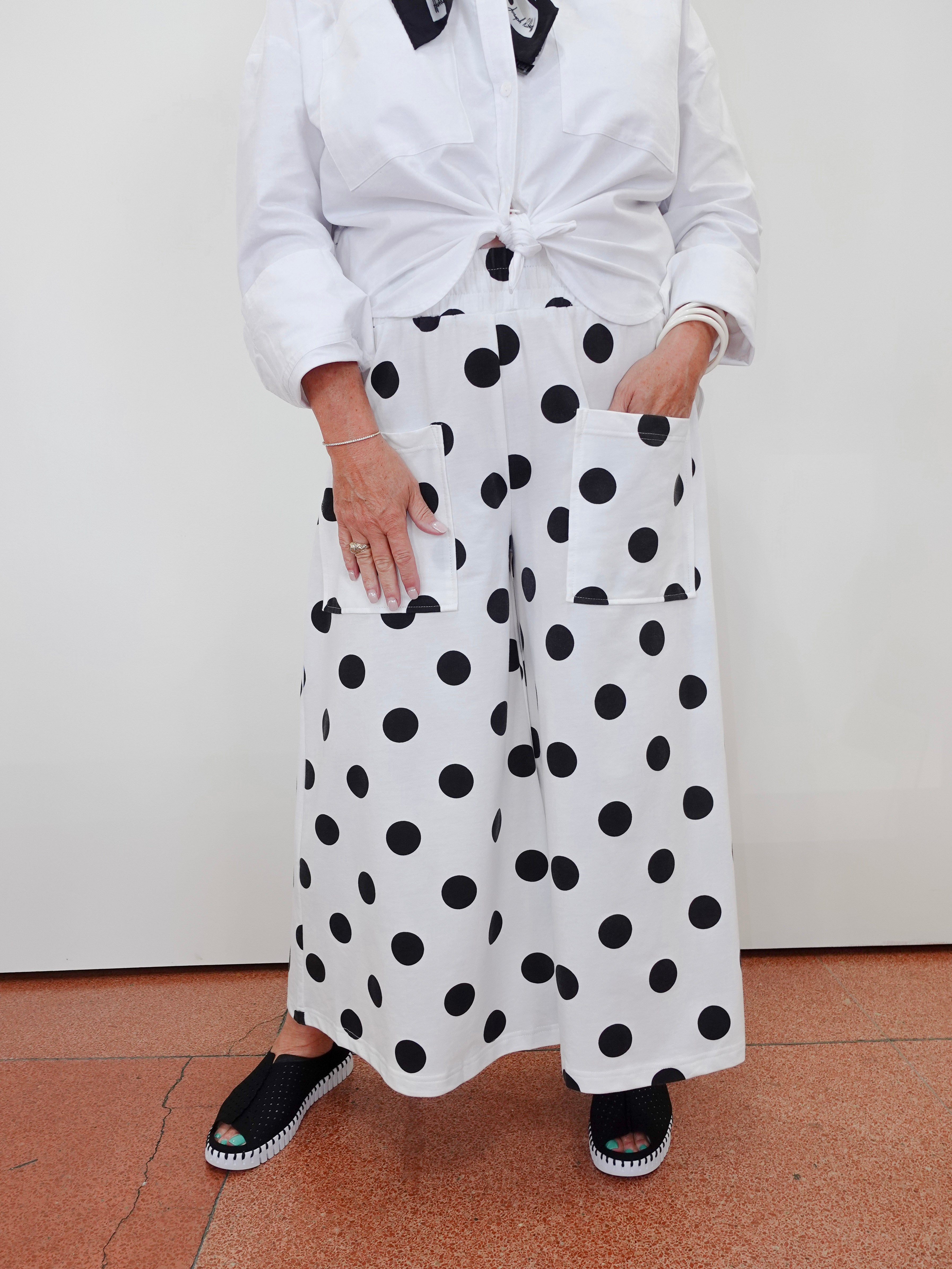 A person wearing white wide-leg pants with large black polka dots, paired with a white tied blouse and black espadrille sandals, standing against a plain off-white wall.