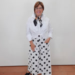 A woman wearing a white long-sleeve button-up shirt tied at the waist, white wide-leg pants with black polka dots, and a black-and-white patterned neck scarf, standing against a plain white wall.