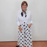A woman wearing a white long-sleeve button-up shirt tied at the waist, white wide-leg pants with black polka dots, and a black-and-white patterned neck scarf, standing against a plain white wall.