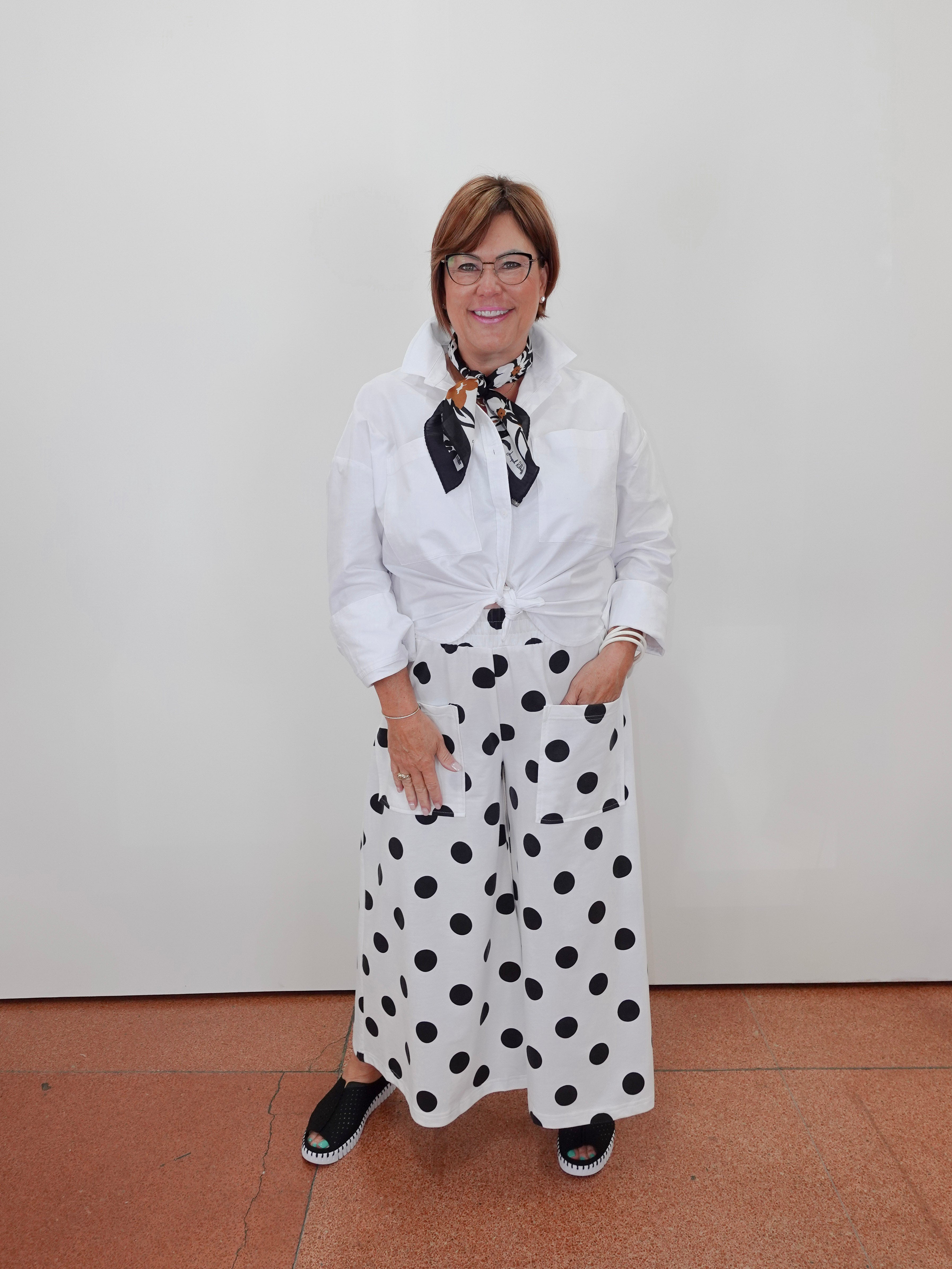 A woman wearing a white long-sleeve button-up shirt tied at the waist, white wide-leg pants with black polka dots, and a black-and-white patterned neck scarf, standing against a plain white wall.