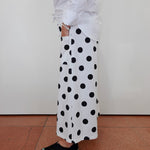 A person wearing a white long-sleeve shirt and white wide-leg pants with large black polka dots, paired with black slip-on shoes, standing against a plain off-white wall on a terracotta tiled floor.