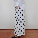 A person wearing a white long-sleeve shirt and white wide-leg pants with large black polka dots, paired with black slip-on shoes, standing against a plain off-white wall on a terracotta tiled floor.