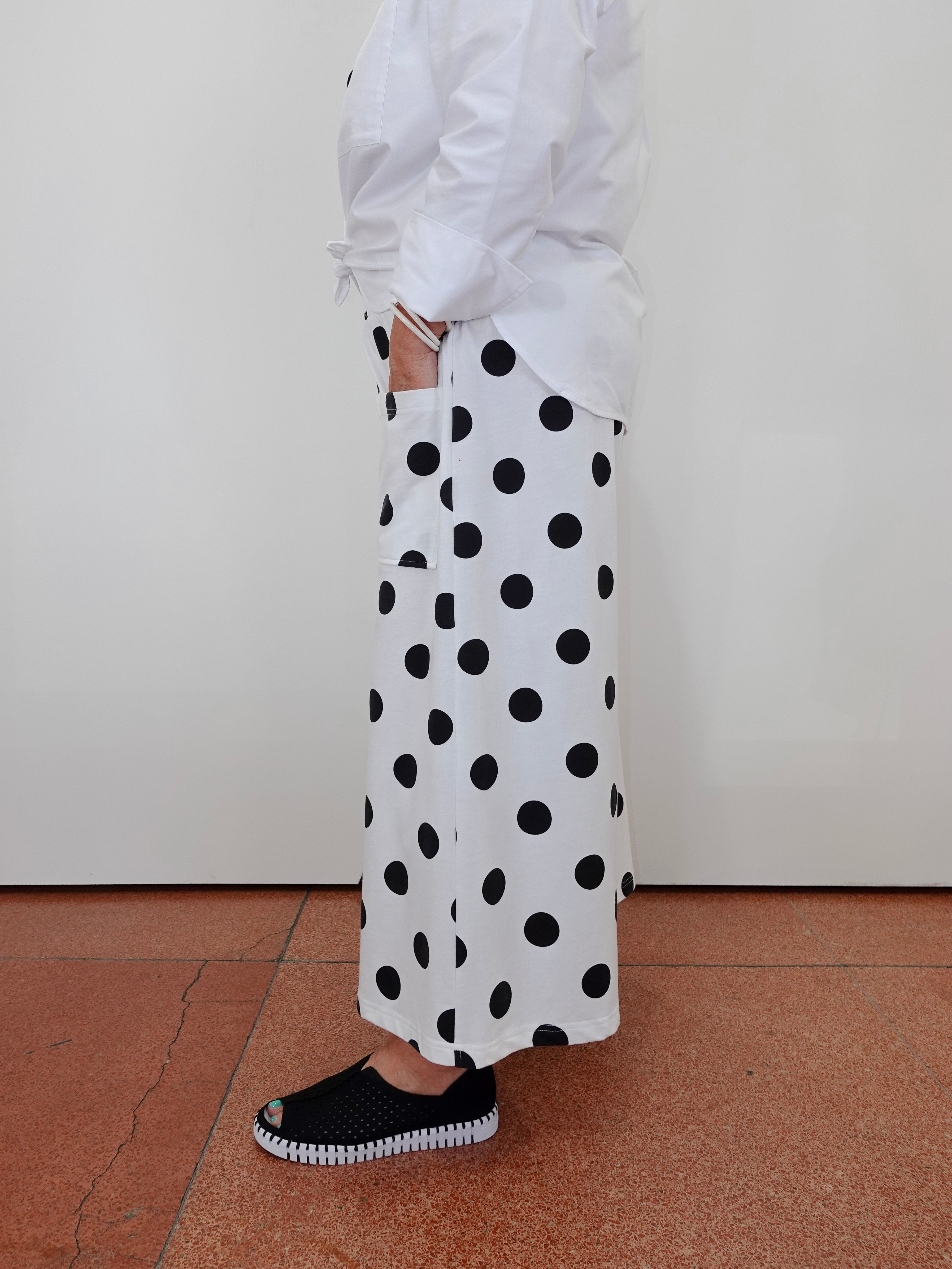 A person wearing a white long-sleeve shirt and white wide-leg pants with large black polka dots, paired with black slip-on shoes, standing against a plain off-white wall on a terracotta tiled floor.