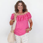 Woman wearing a pink blouse with ruffles and white floral pants, holding a beige handbag on a white background.
