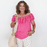 Woman wearing a pink blouse with ruffles and white floral pants, holding a beige handbag on a white background.