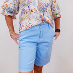 Person wearing a colorful floral blouse and light blue shorts on a plain background