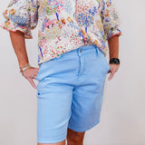 Person wearing a colorful floral blouse and light blue shorts on a plain background