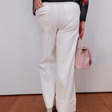 Person wearing white pants and a floral sweater, holding a pink handbag against a plain background