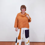 Woman wearing an orange top and patterned pants against a white wall.