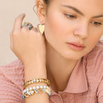 Woman wearing gold jewelry including earrings, rings, and bracelets on a beige background