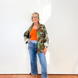 Person wearing a camouflage jacket, orange top, and blue jeans against a white wall.
