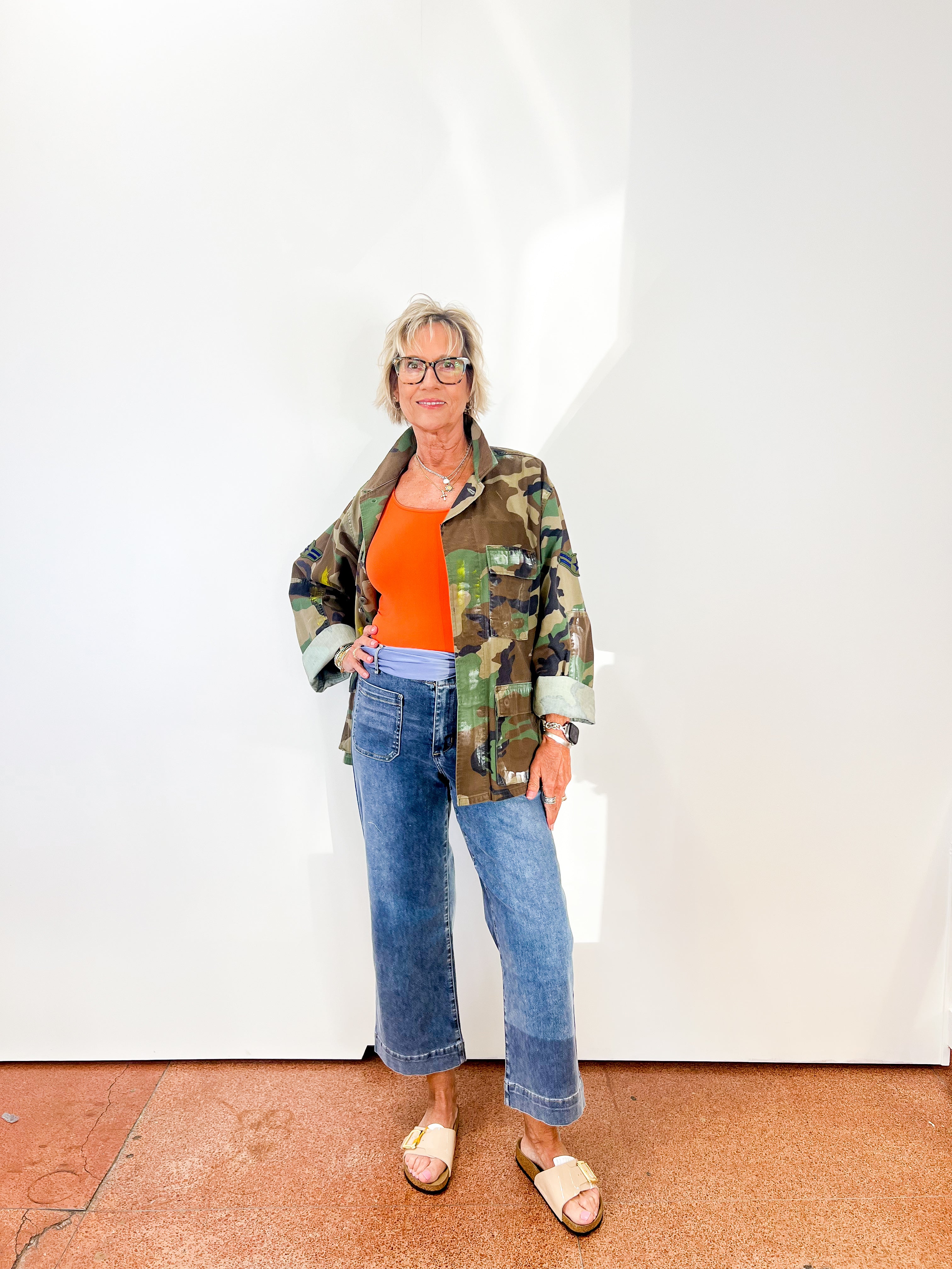 Person wearing a camouflage jacket, orange top, and blue jeans against a white wall.
