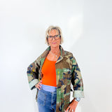 Woman wearing a camouflage jacket over an orange shirt with blue jeans on a white background