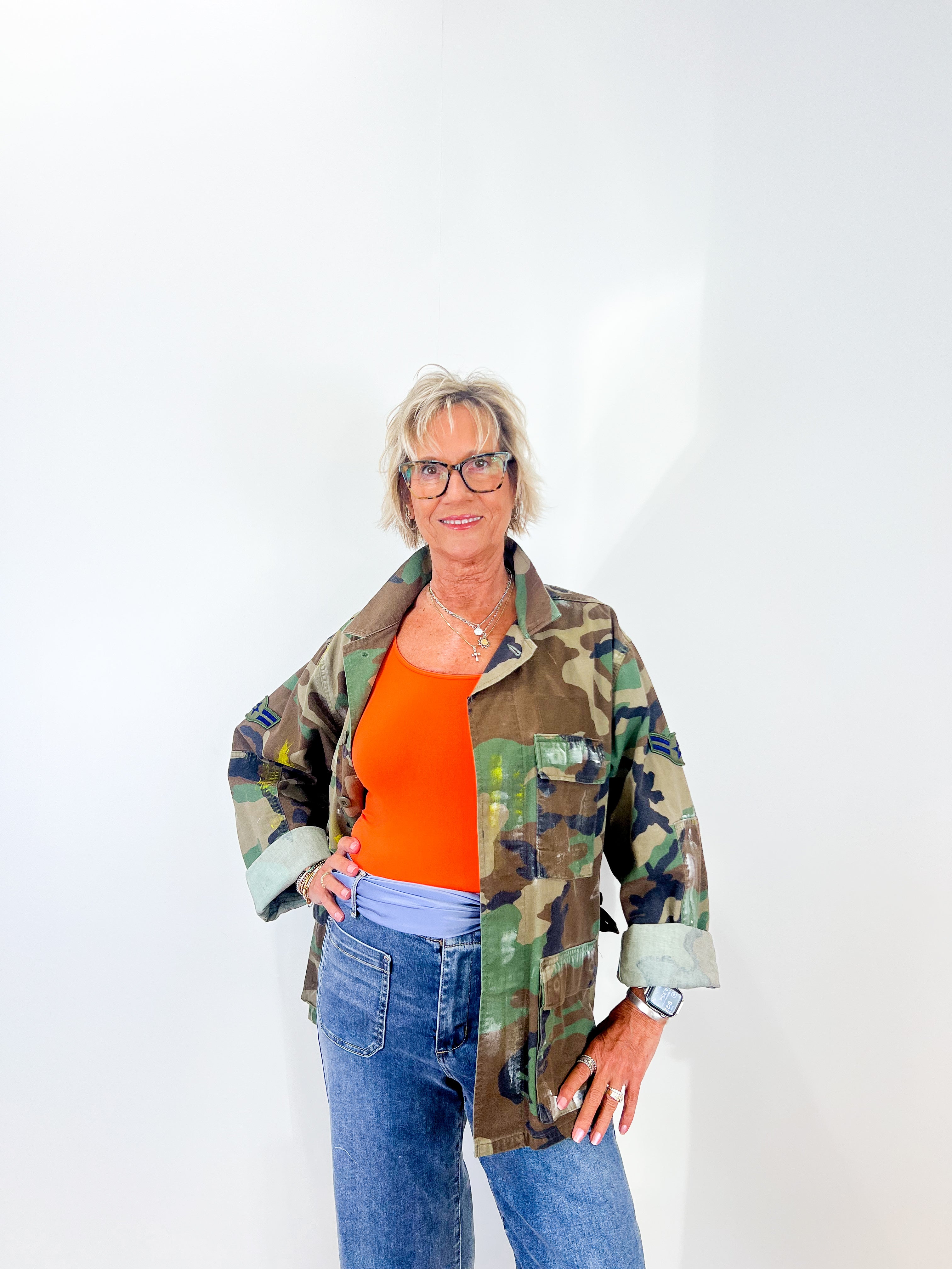 Woman wearing a camouflage jacket over an orange shirt with blue jeans on a white background