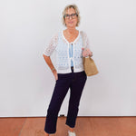 Woman wearing a white lace top, navy pants, and glasses against a plain background