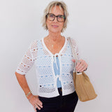 Woman wearing a white lace top and black pants, holding a beige handbag against a plain background