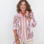 Woman wearing a pink and white patterned blouse with beige shorts, holding a pink handbag on a white background.