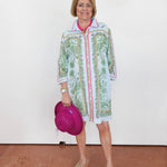 Woman wearing a colorful patterned shirt over a white dress, holding a pink hat against a white background.