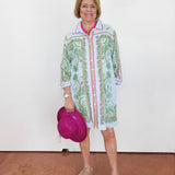 Woman wearing a colorful patterned shirt over a white dress, holding a pink hat against a white background.