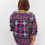 A woman viewed from behind wearing a colorful, patterned long-sleeve blouse with blue jeans, standing against a plain white studio background.