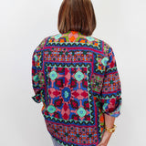 A woman viewed from behind wearing a colorful, patterned long-sleeve blouse with blue jeans, standing against a plain white studio background.