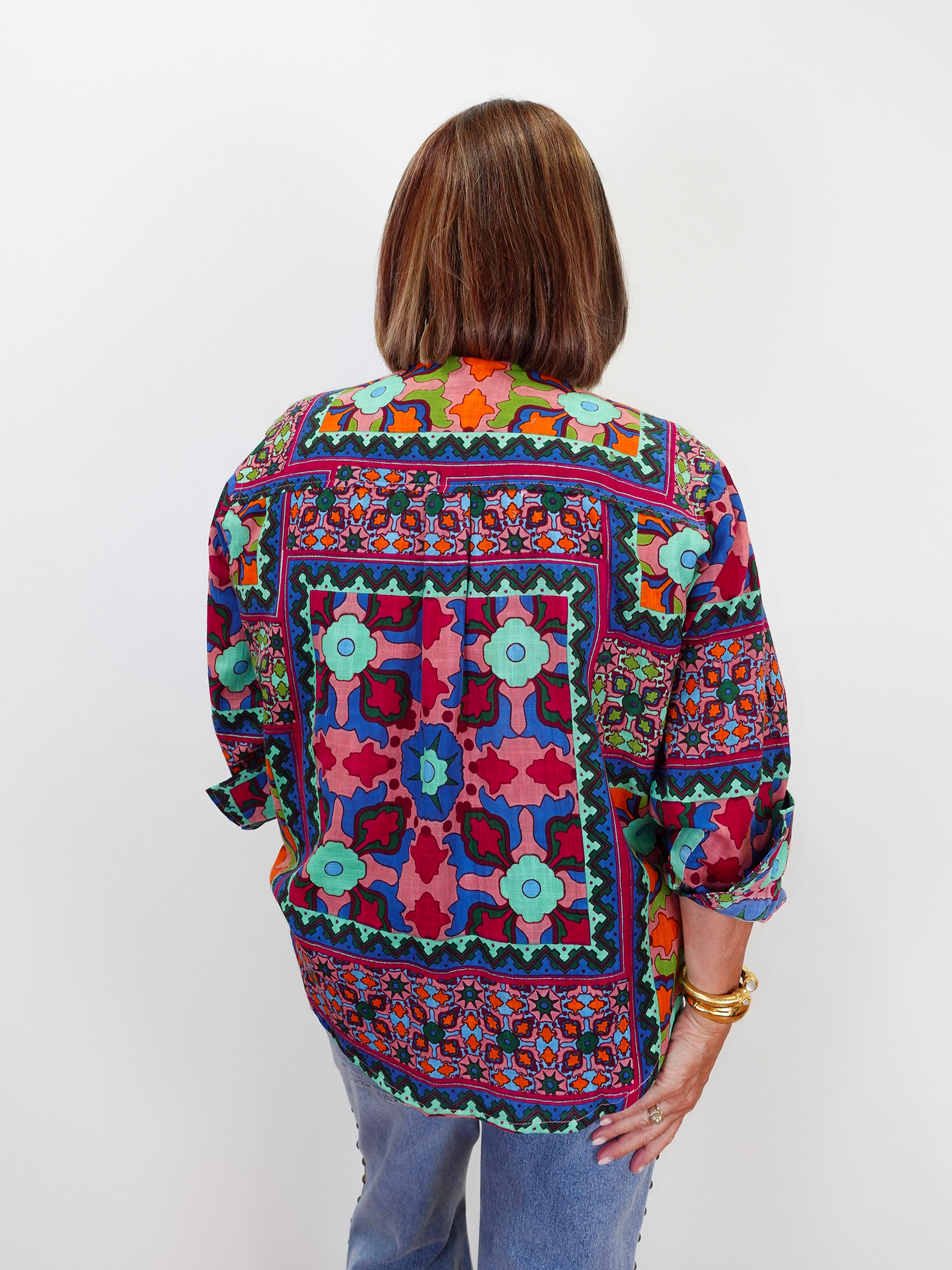 A woman viewed from behind wearing a colorful, patterned long-sleeve blouse with blue jeans, standing against a plain white studio background.